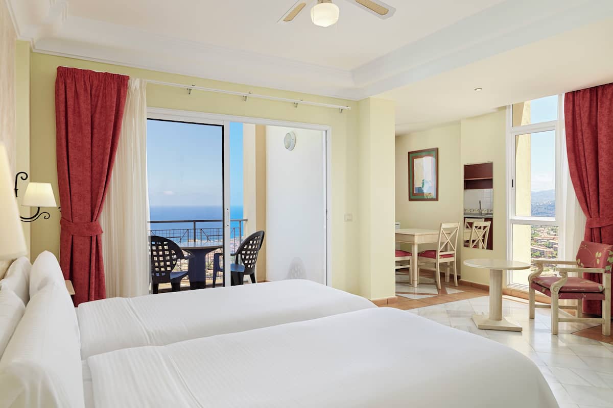 A hotel room with two beds, a balcony with a table and chairs, a kitchenette, and a view of the sea.