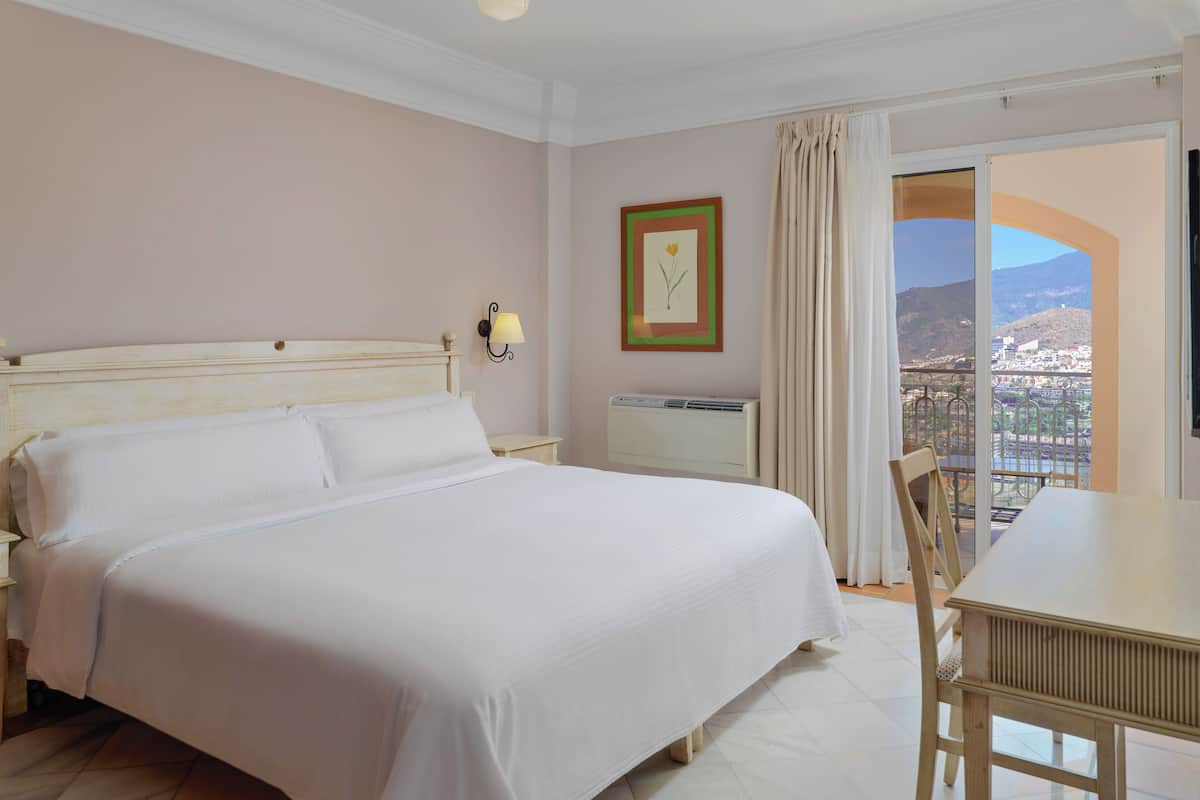 A hotel room with a large bed, a bedside table, a lamp, a framed picture on the wall, and a balcony with a view.