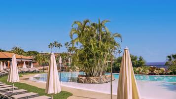 Zwembad van een resort met ligbedden en parasols, omringd door palmbomen en een helder uitzicht op de oceaan.