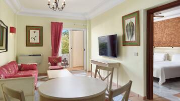A hotel room with a bed, a TV, a dining table with chairs, a sofa, and a balcony with a view of greenery.
