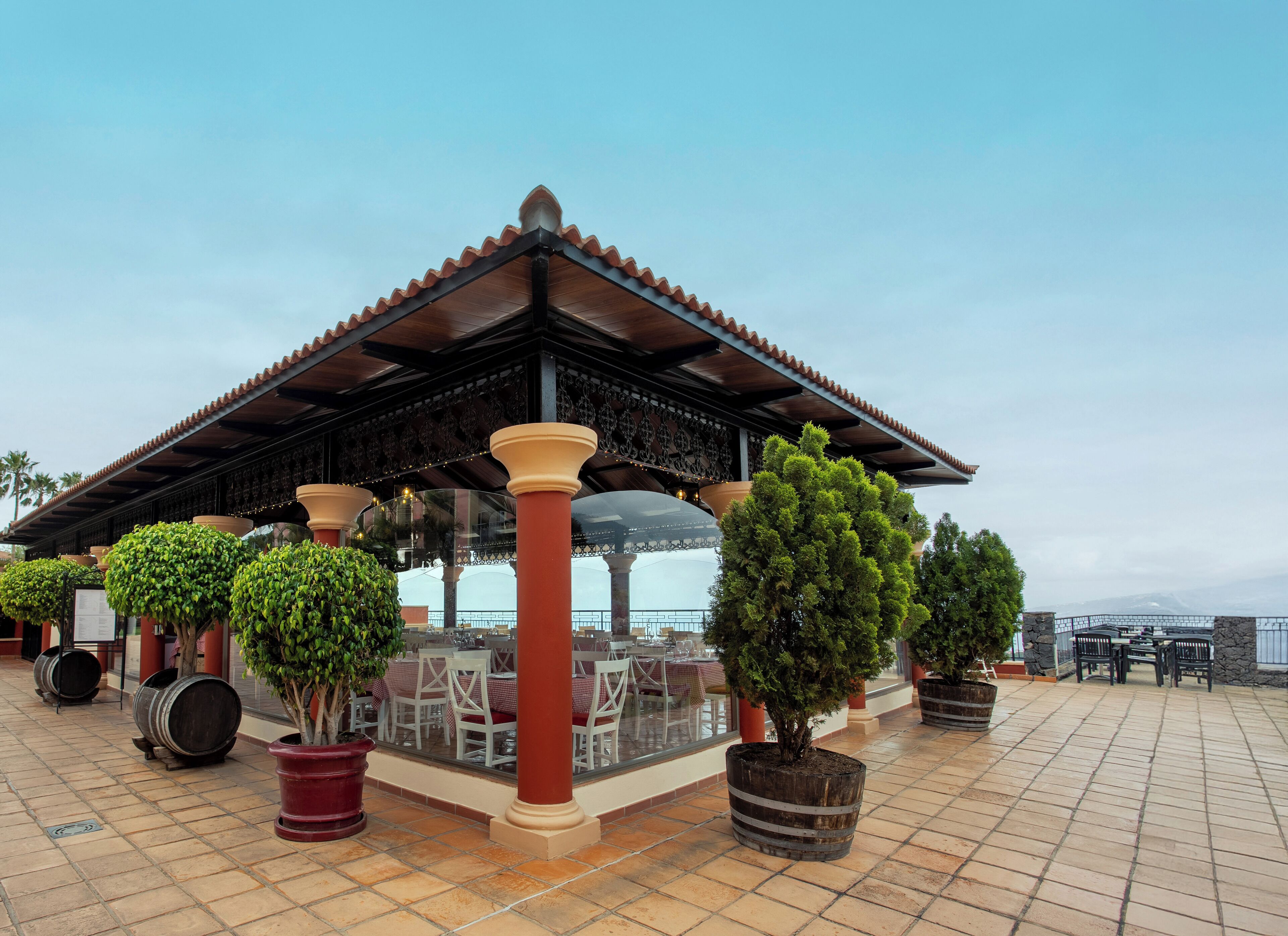 A covered outdoor seating area with tables and chairs, surrounded by potted plants and a pergola structure.
