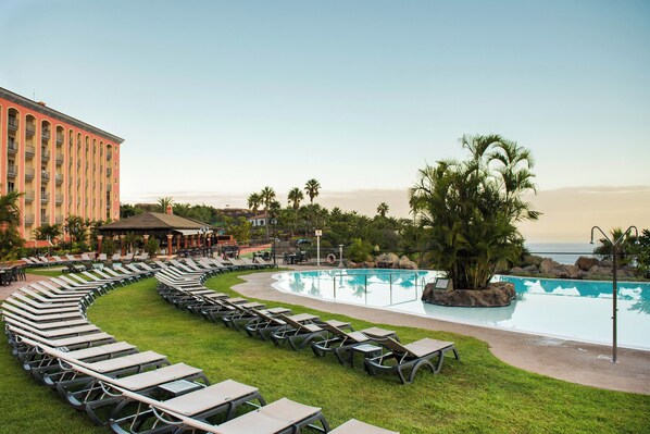 Zona de piscina en un resort, con sillones de descanso y un edificio al fondo.