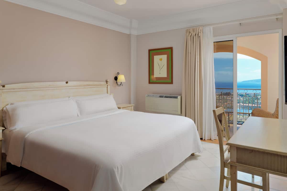 A hotel room with a large bed, a small table, a chair, a wall-mounted air conditioner, a nightstand, a lamp, and a window with a view of the sea.