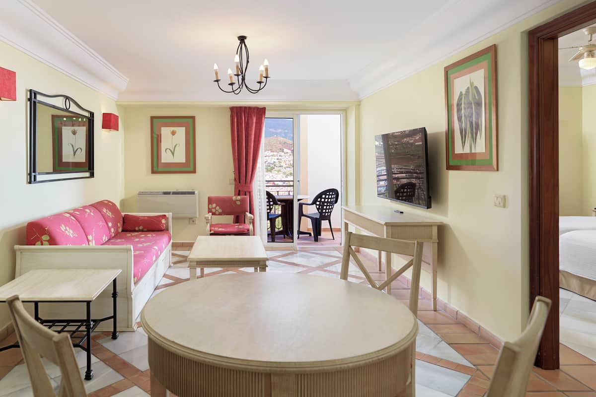 A room with a round table, a sofa, a TV, and a balcony with chairs.