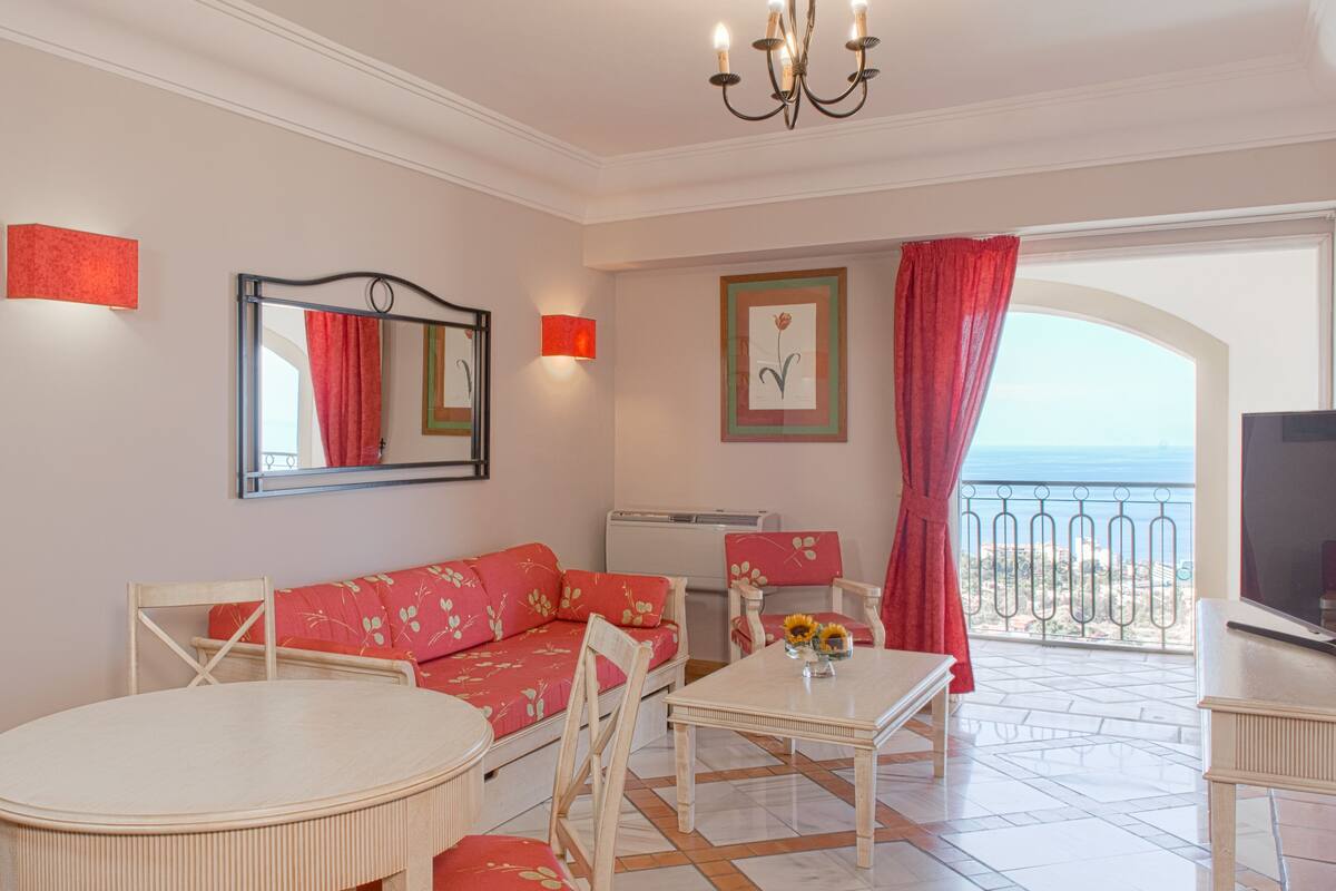 A cozy living room with a sofa, chairs, a dining table, and a TV. There is a balcony with a view of the sea.