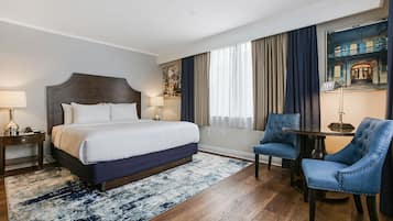 A hotel room with a large bed, two bedside tables with lamps, a blue armchair, a round table, and a large window with curtains.