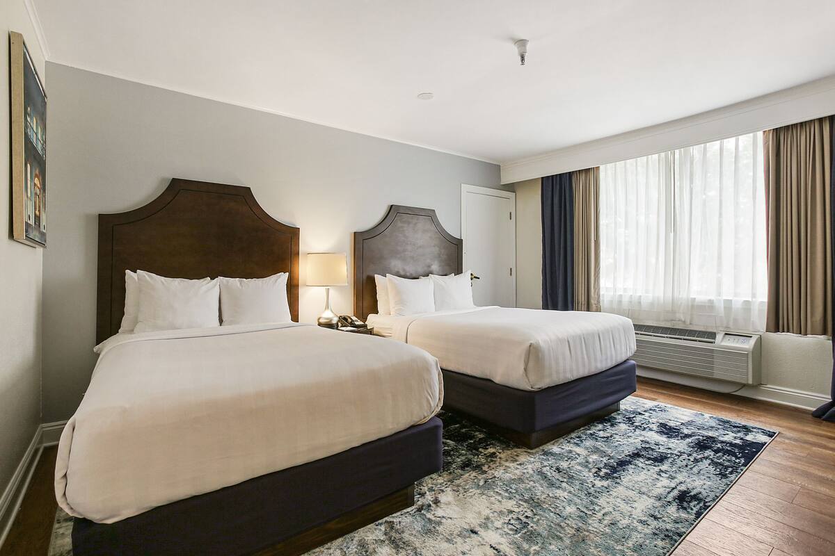 Une chambre d’hôtel avec deux lits, une grande fenêtre avec des rideaux, un climatiseur fixé au mur et un tapis à motifs.