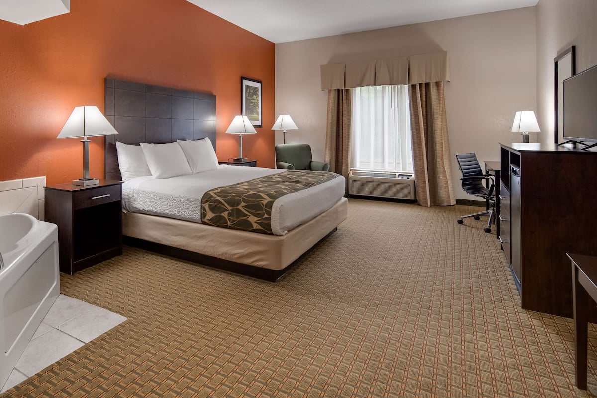 A hotel room with a large bed, a separate desk, a chair, a desk lamp, a TV, a bathroom with a bathtub, and a window with curtains.