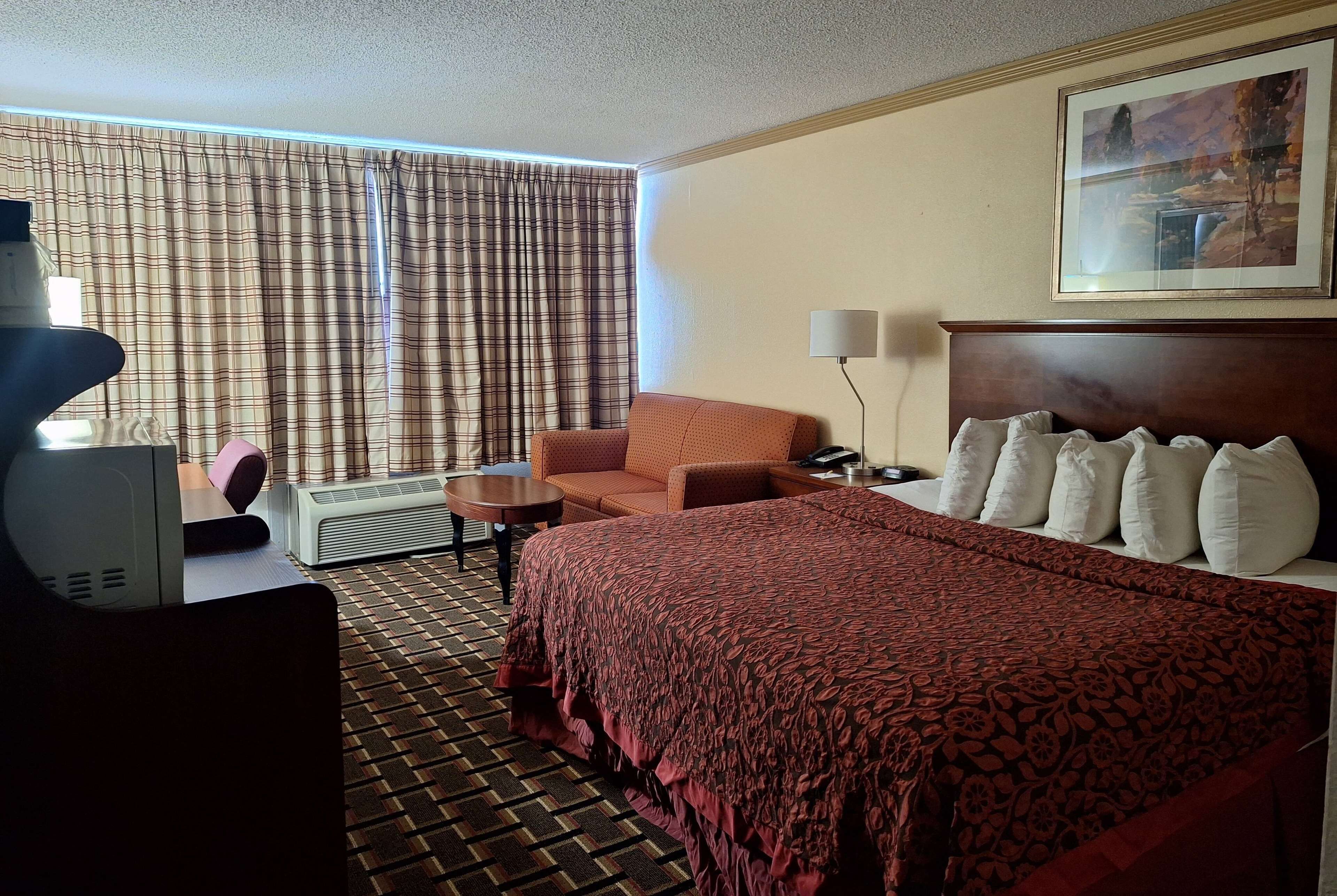 A hotel room with a large bed, a sofa, a desk with a lamp, a heater, and a painting on the wall.