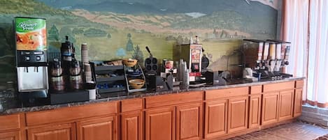 A hotel bar area with a mountain mural, a juice dispenser, coffee machines, and a variety of condiments and utensils on the counter.