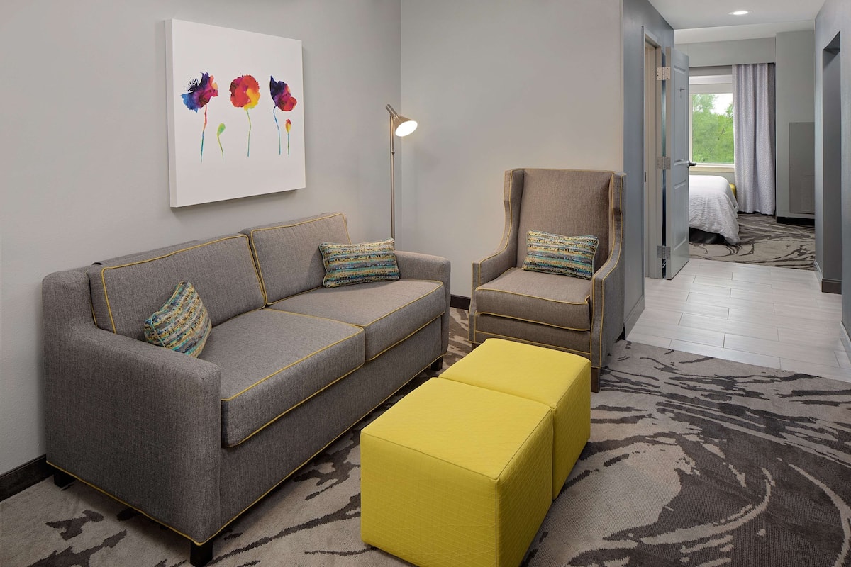 A modern hotel room with a grey sofa, a yellow ottoman, a beige armchair, and a floor lamp. There is a painting on the wall and a window with curtains.