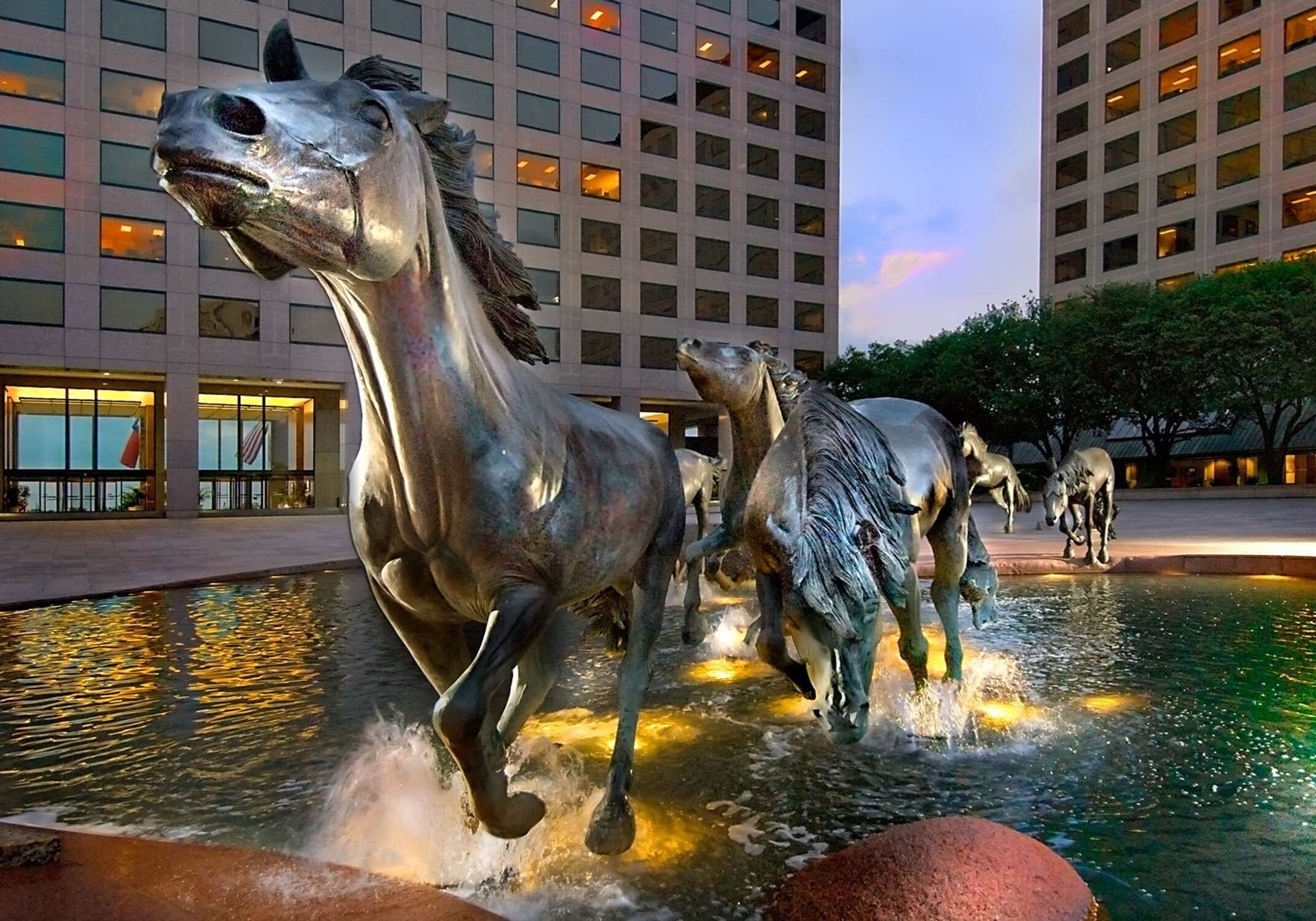 A fountain with bronze horse sculptures in an urban setting with modern buildings in the background.