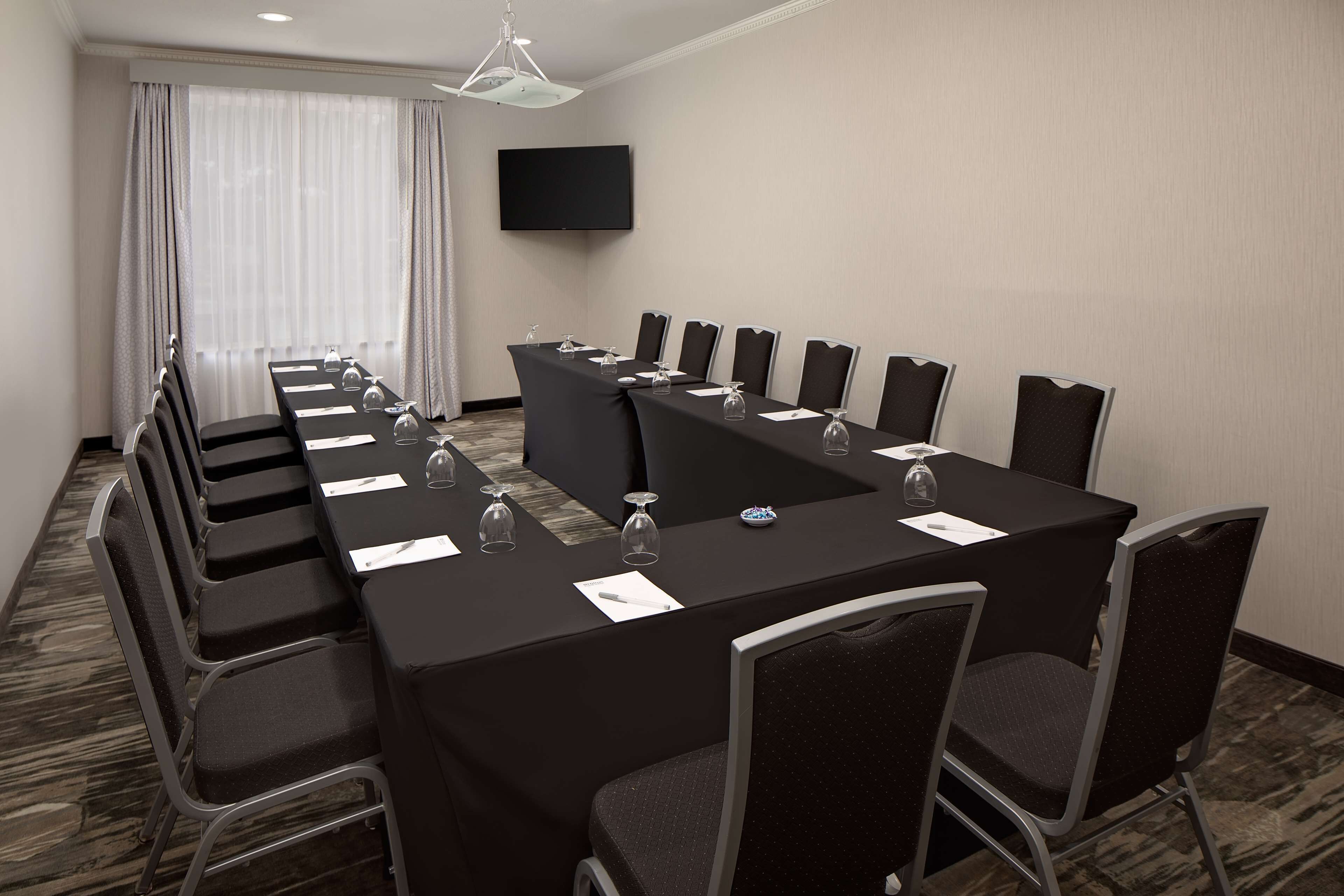 A conference room with a long table, chairs, a flat-screen TV, and a window with curtains.