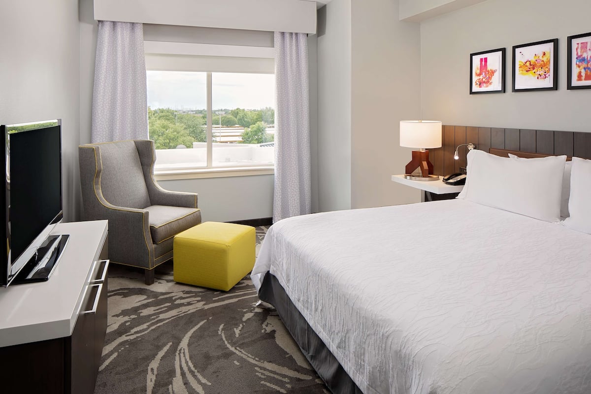 A hotel room with a bed, a desk with a TV, a chair, a yellow ottoman, a nightstand with a lamp, and a window with a view of greenery.