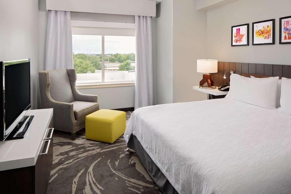 A hotel room with a bed, a desk with a TV, a chair, a yellow ottoman, a nightstand with a lamp, and a window with a view.