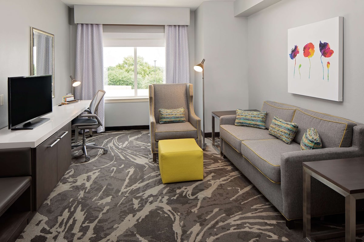 A hotel room with a desk, chair, sofa, ottoman, and artwork on the wall.