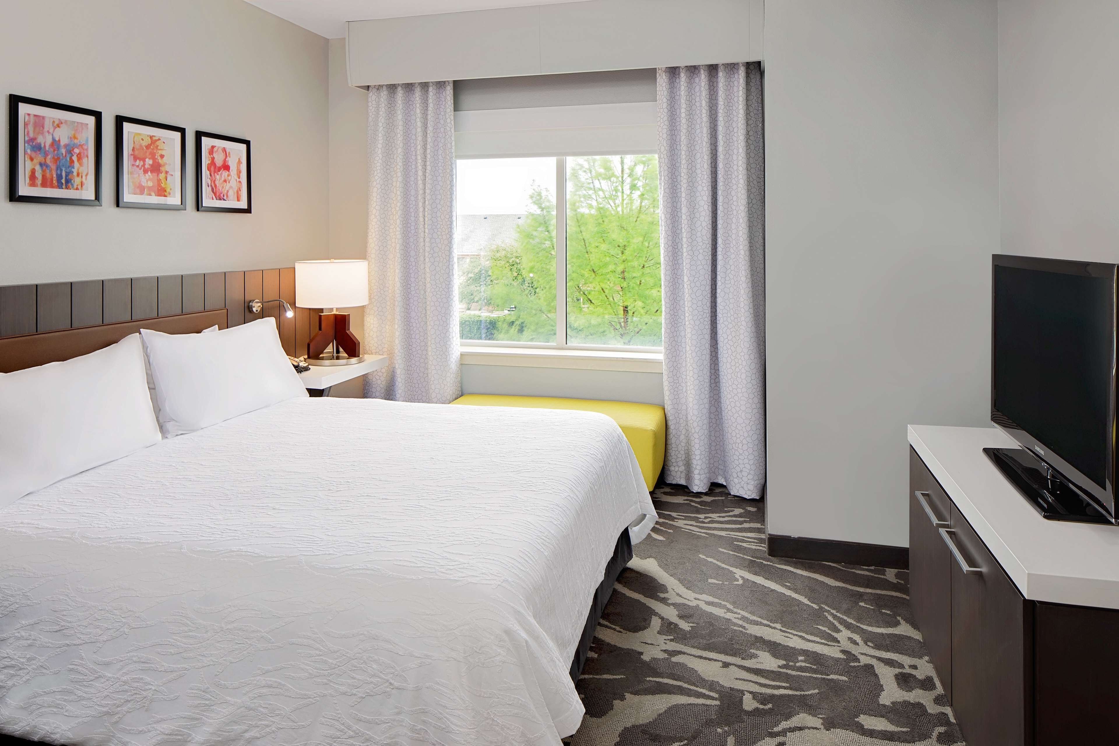 A hotel room with a large bed, a desk with a TV, a window with curtains, and artwork on the wall.