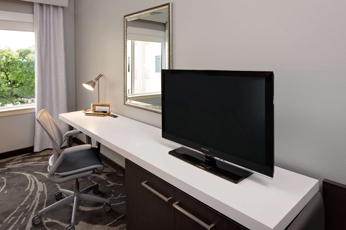 A hotel room with a desk, a flat-screen TV, a chair, a lamp, and a window with a view of trees.