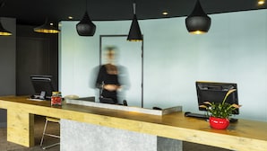 A reception desk with a wooden top and a concrete base, a computer monitor, a phone, and a red vase with a plant. - ibis Tarbes Odos (Odos)