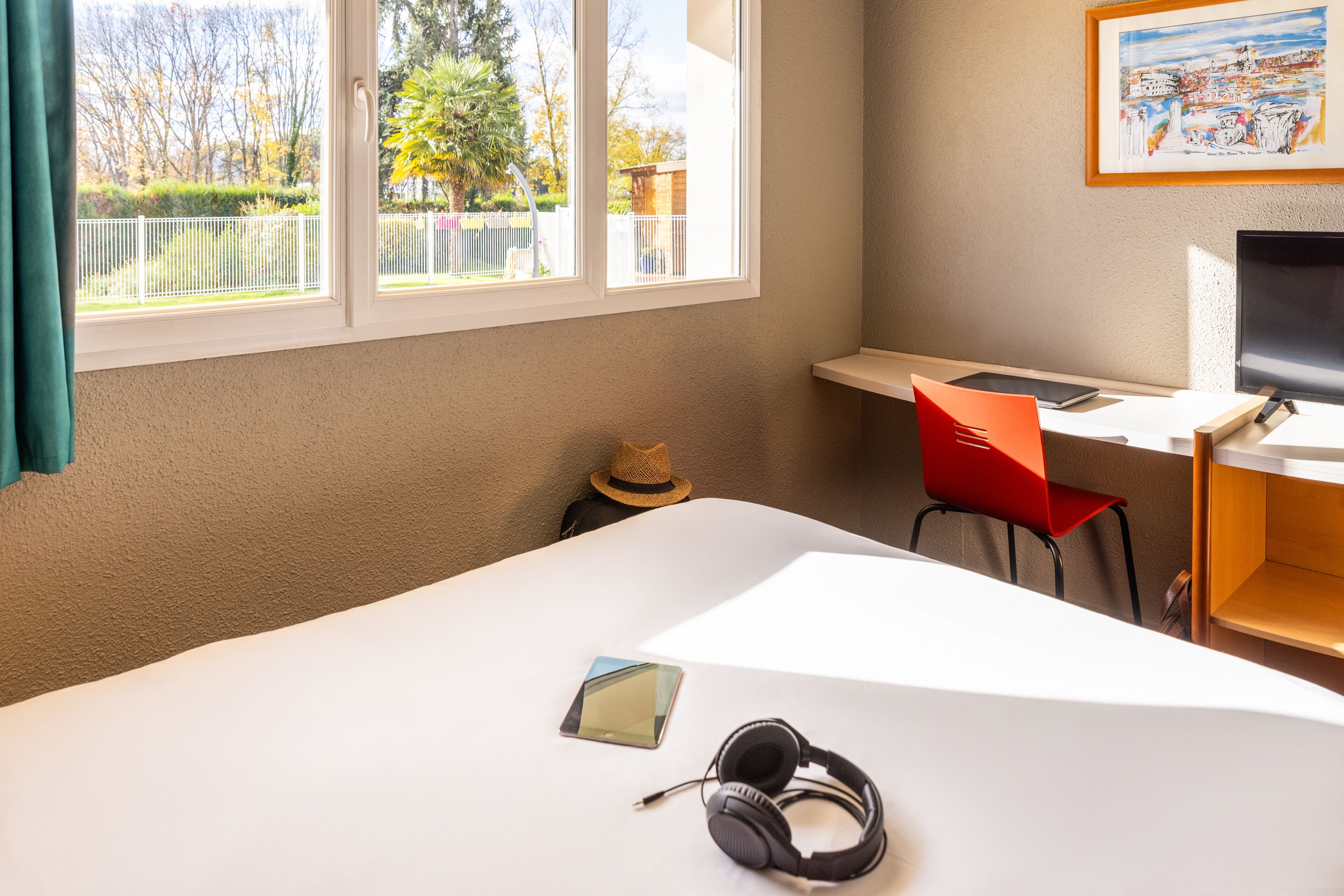 Een kamer met een bed, een bureau met een computer, een stoel en een raam met uitzicht op bomen en een hek.