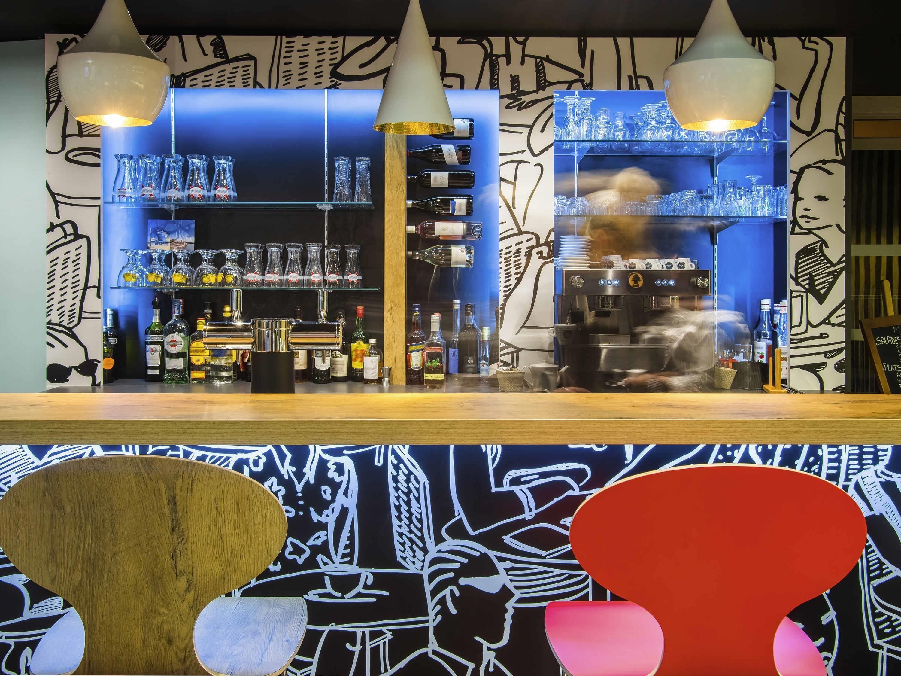 A bar area with shelves of bottles and glasses, a coffee machine, and a person working behind the counter.