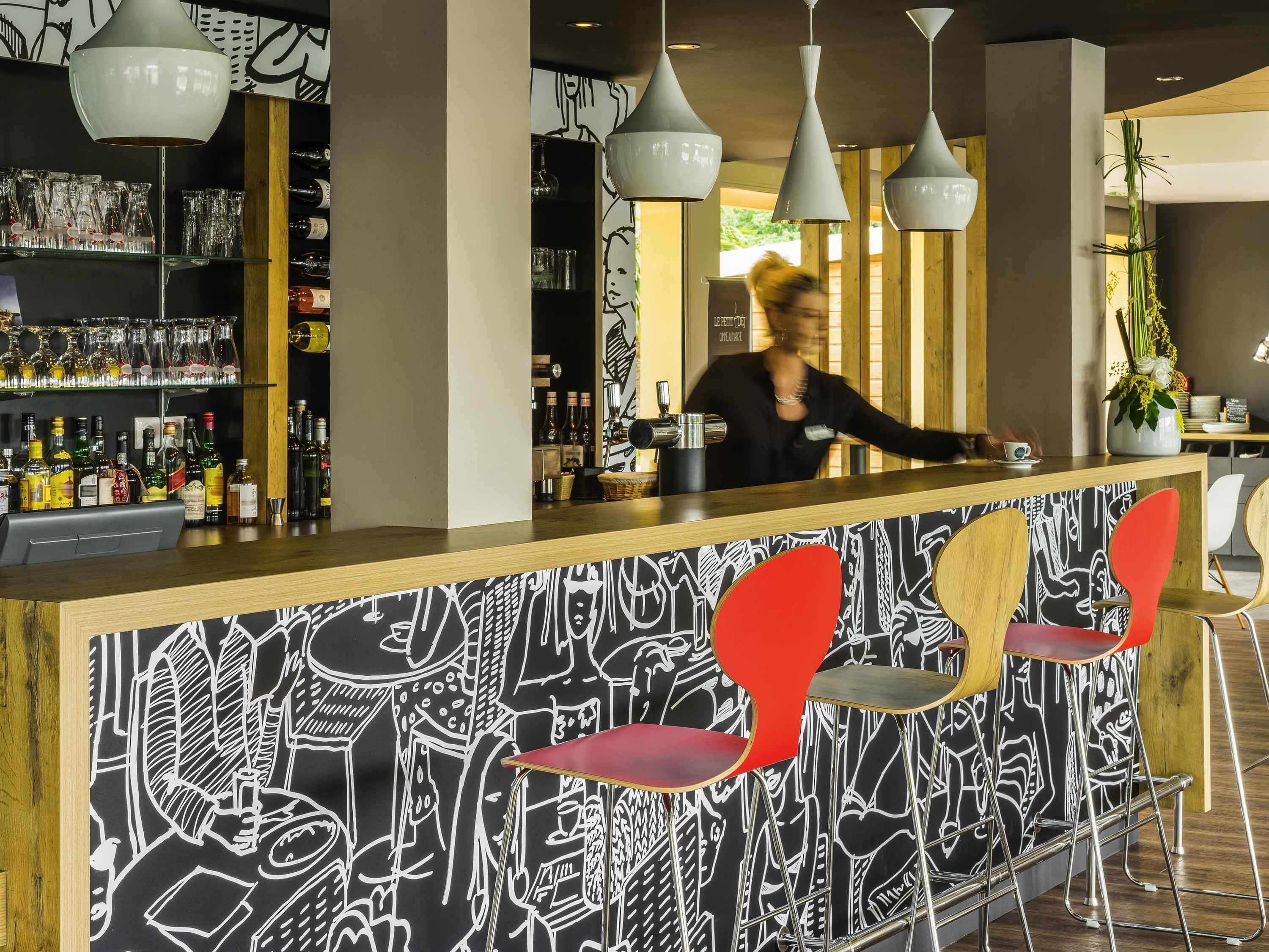Zona de bar com balcão, prateleiras com garrafas e um barman.