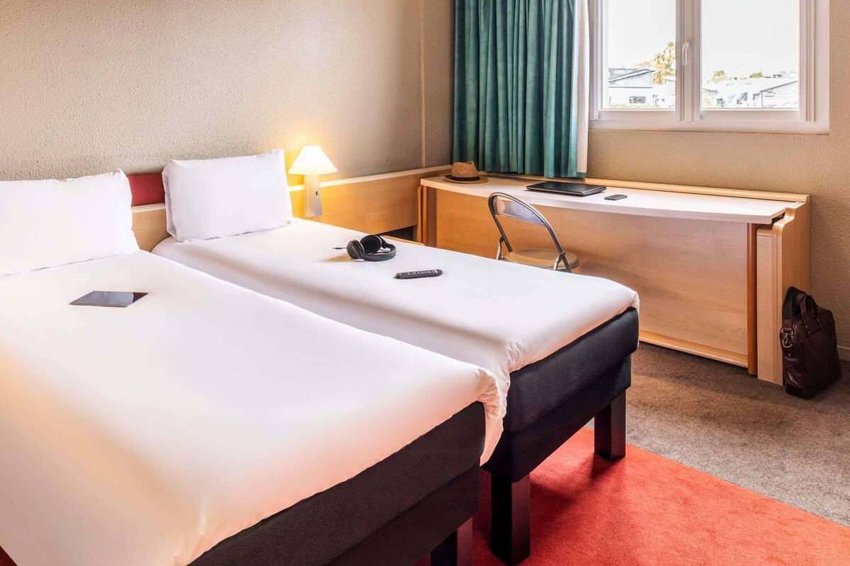 A hotel room with two beds, a desk, a chair, a lamp, and a view of the outside.