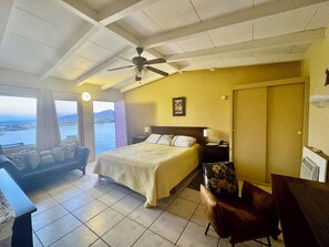 A bedroom with a bed, a desk, a chair, a ceiling fan, and a view of the sea.