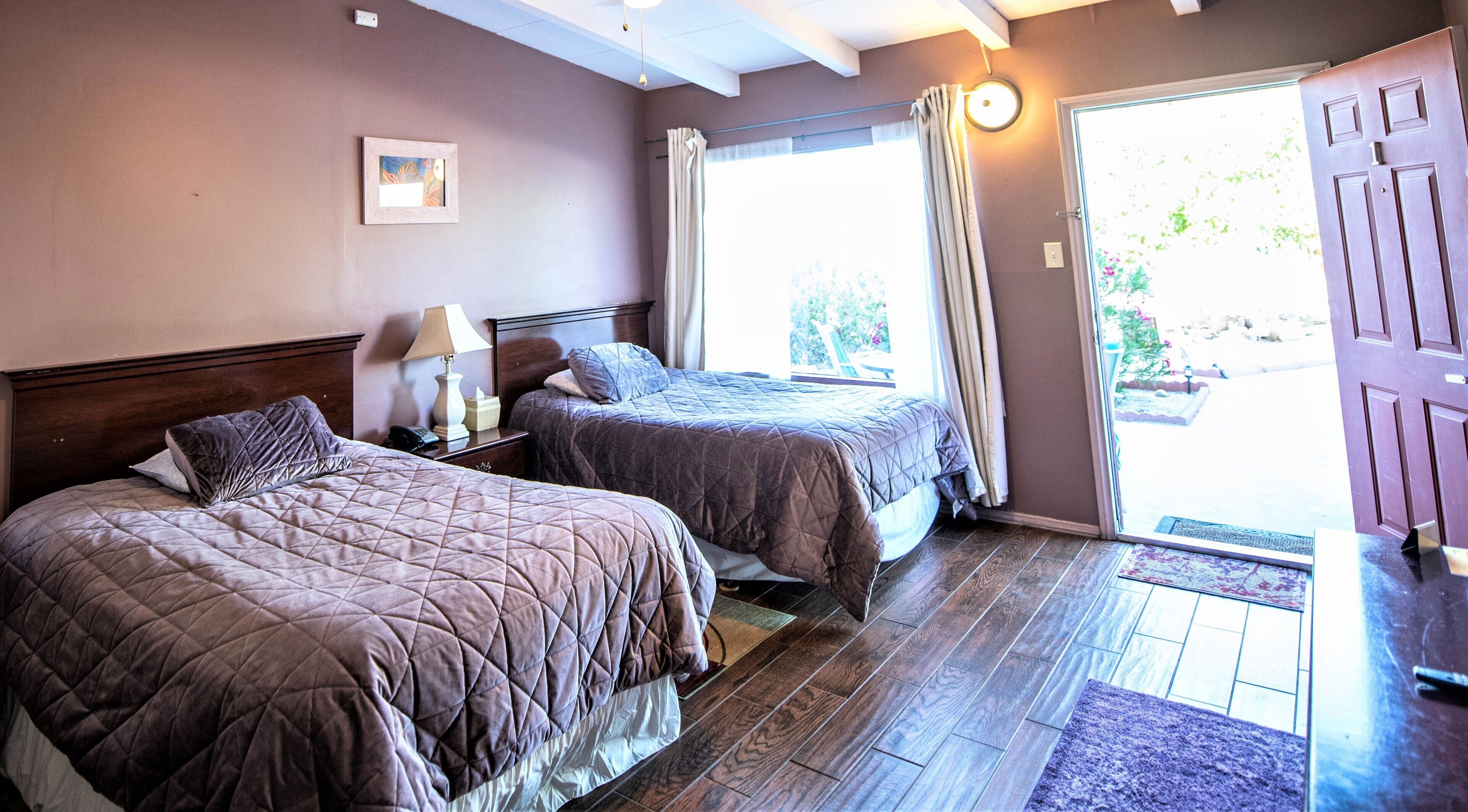 A hotel room with two beds, a nightstand with a lamp, a framed picture on the wall, and a door leading outside.