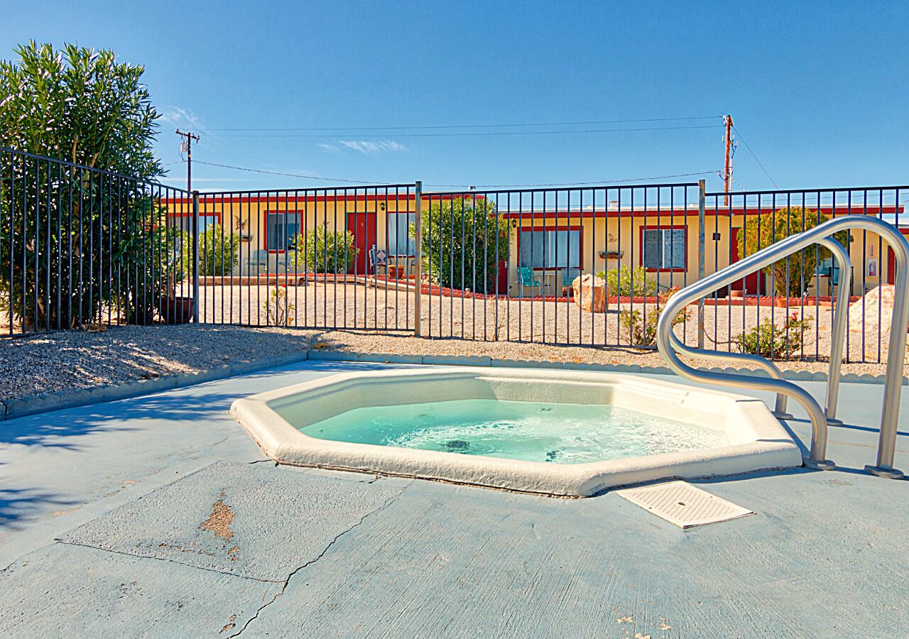 Une piscine avec un escalier en métal, entourée d’une clôture, et un bâtiment avec une porte rouge et des fenêtres en arrière-plan.