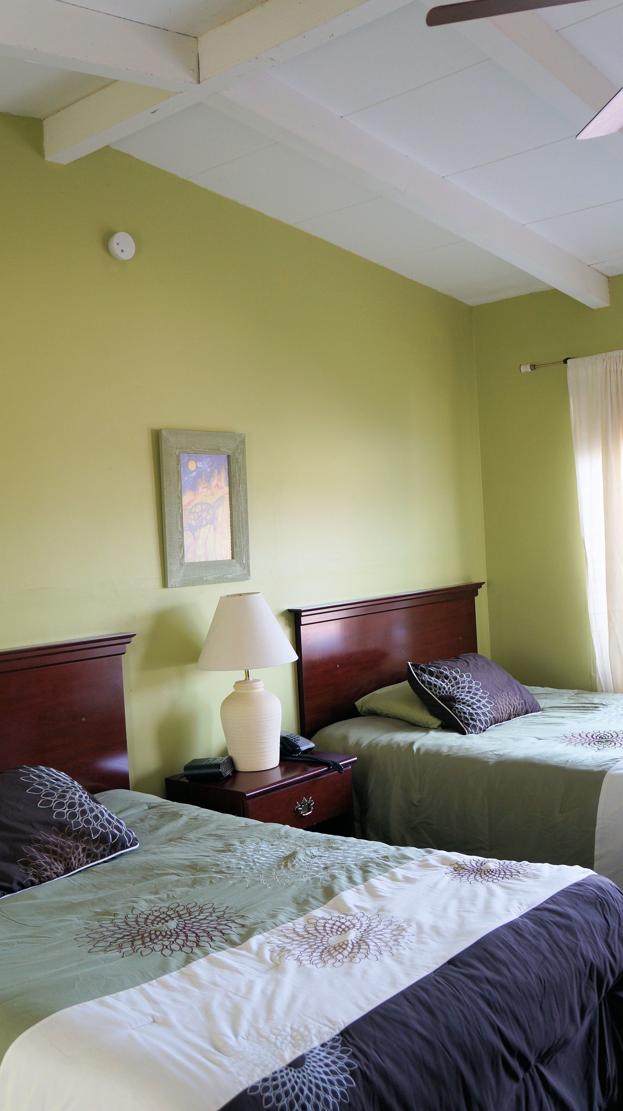 A hotel room with two beds, a nightstand with a lamp, a framed picture on the wall, and a ceiling fan.