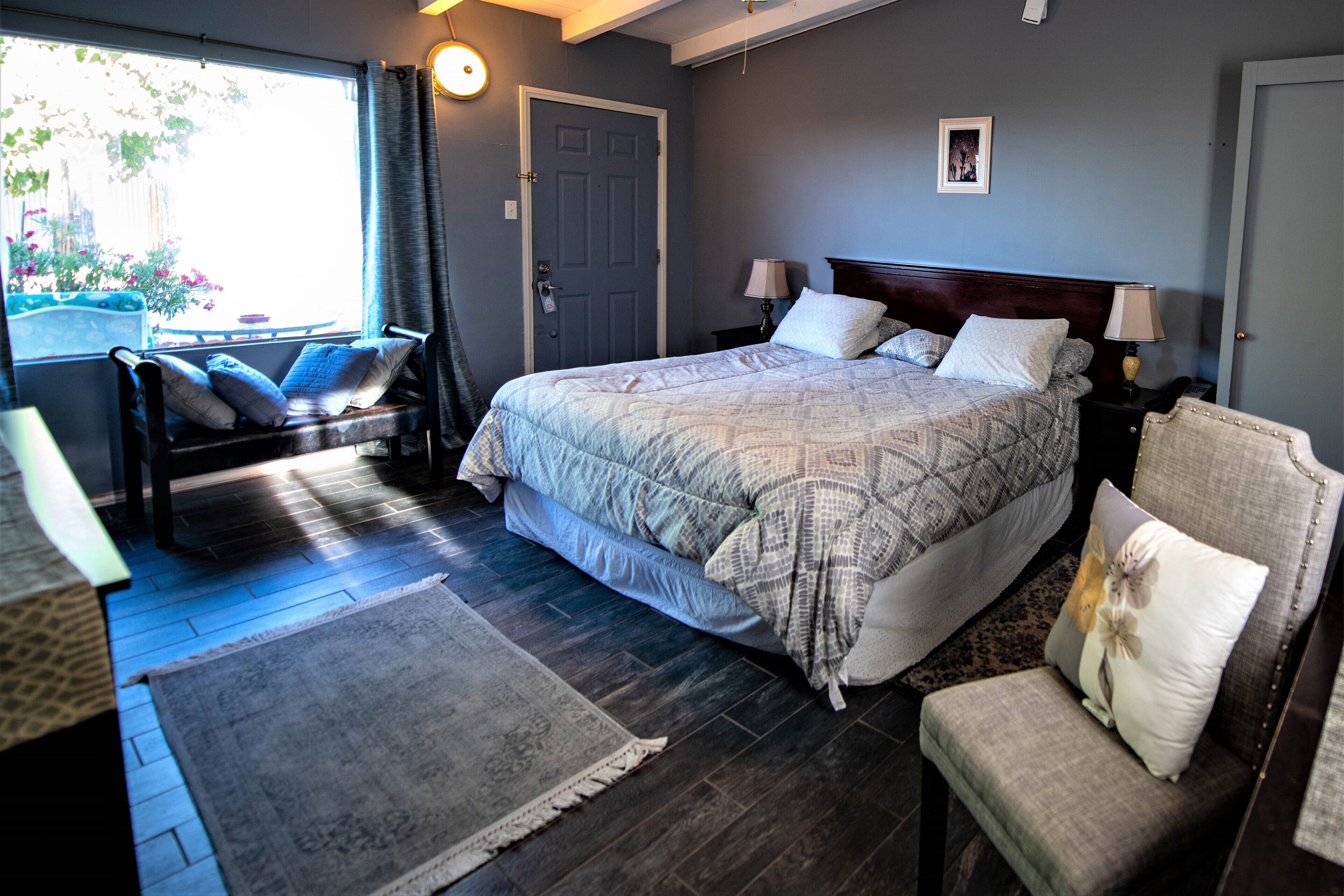Un dormitorio con una cama grande, dos mesitas de noche con lámparas, una silla, un ventanal con cortinas y una puerta.