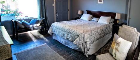 Un dormitorio con una cama grande, dos mesitas de noche con lámparas, una silla, un ventanal con cortinas y una puerta.