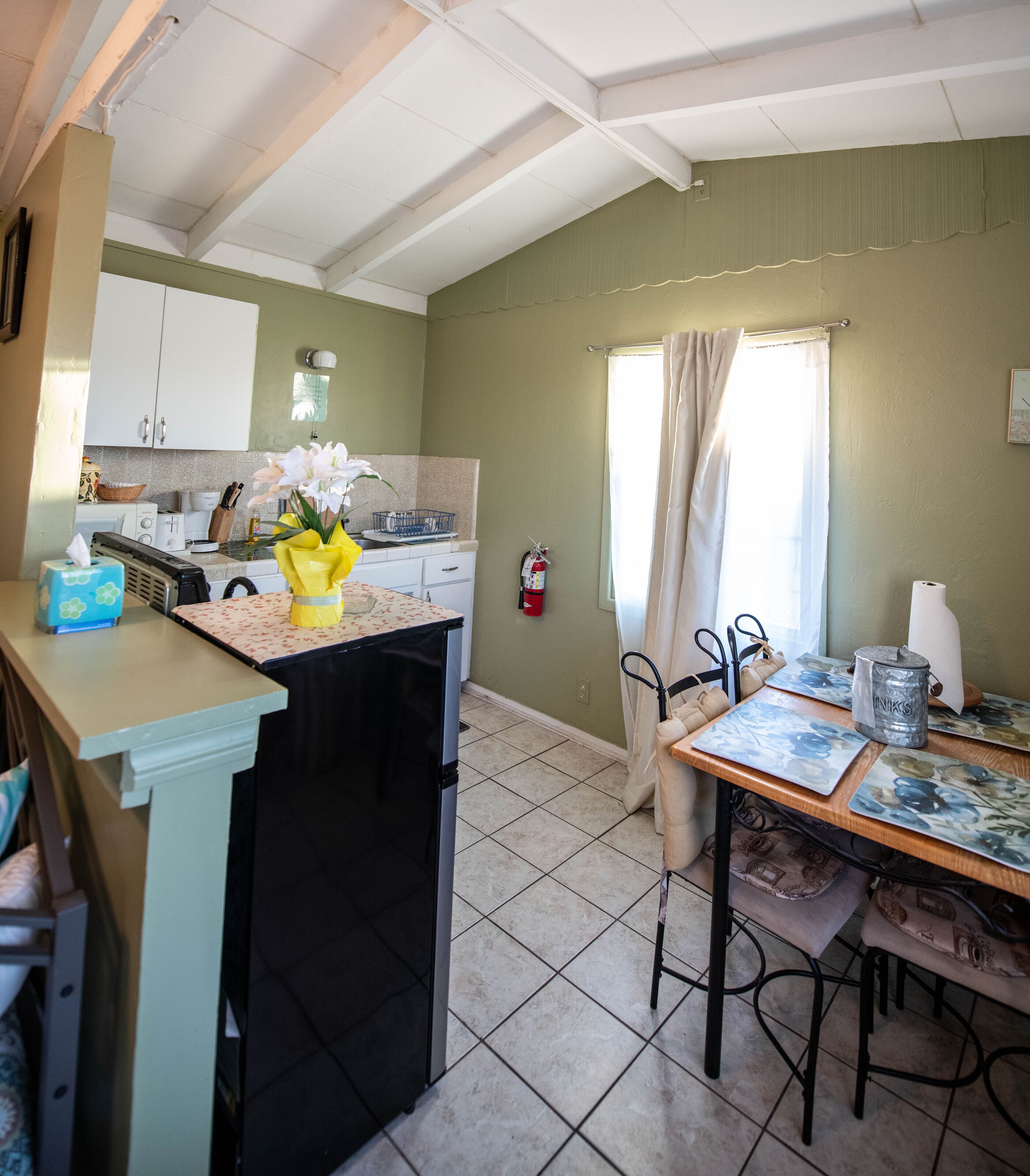 A kitchen with white cabinets, a black refrigerator, a dining table with chairs, a window with curtains, and a fire extinguisher on the wall.