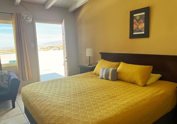 A bed with a patterned bedspread, a nightstand with a lamp, a framed picture on the wall, and a view through an open door. - Harmony Motel (Twentynine Palms)