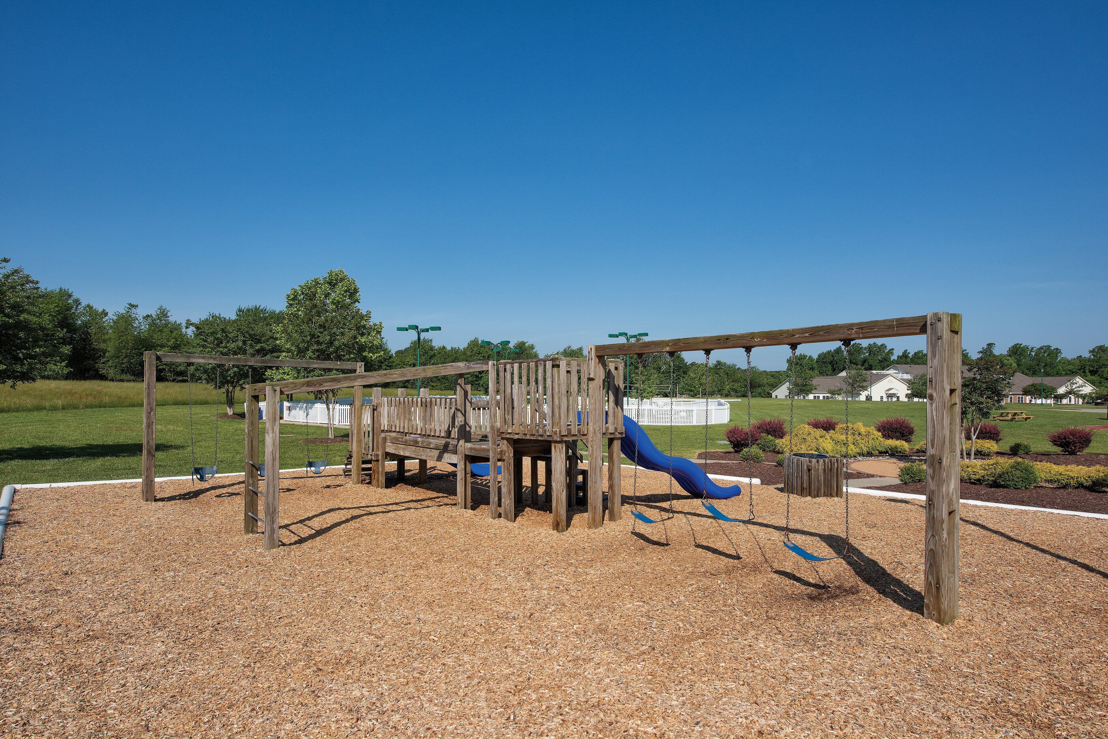 A wooden playground structure with a slide and swings, surrounded by a grassy area and a clear sky.