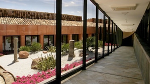 A corridor with a view of a building with a tiled roof and a garden area with rocks and pink flowers. - Hotel Cigarral El Bosque (Toledo)