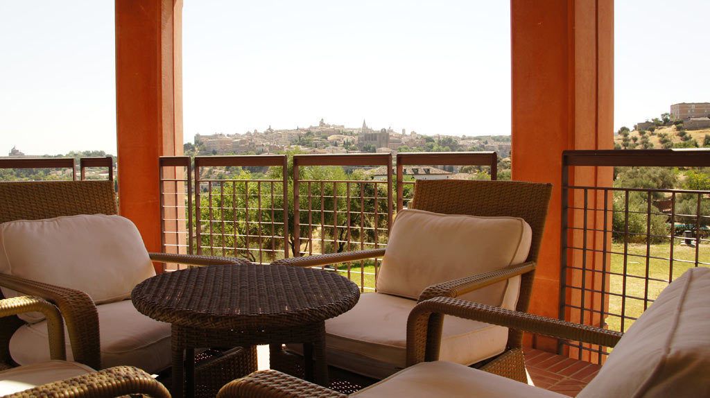 A balcony with wicker furniture, including two chairs and a small table, overlooking a cityscape.