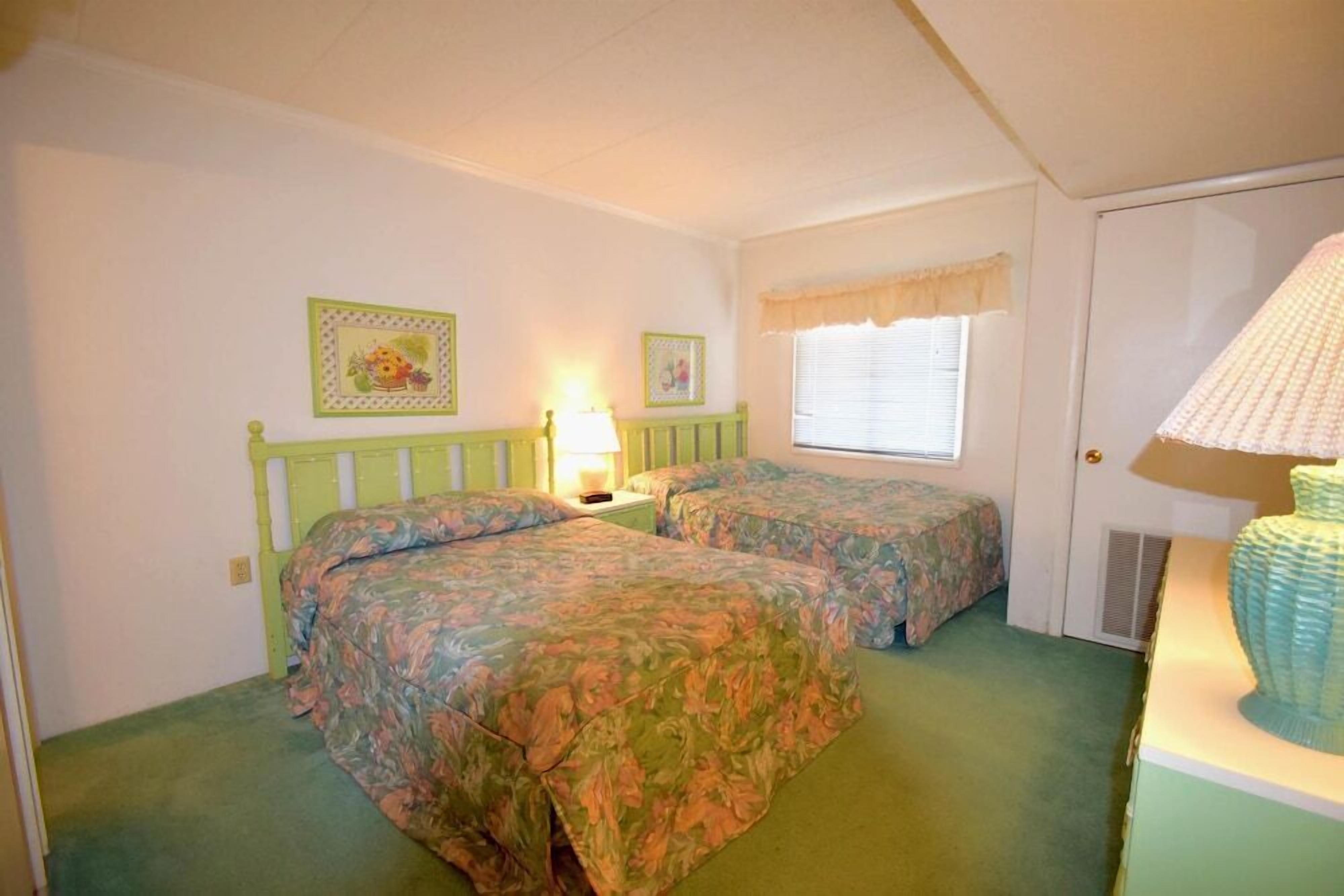 Une chambre avec deux lits, une fenêtre avec des stores, une lampe et une porte.