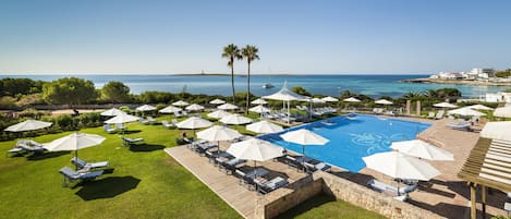 A pool area with lounge chairs and umbrellas, surrounded by greenery and a view of the sea.