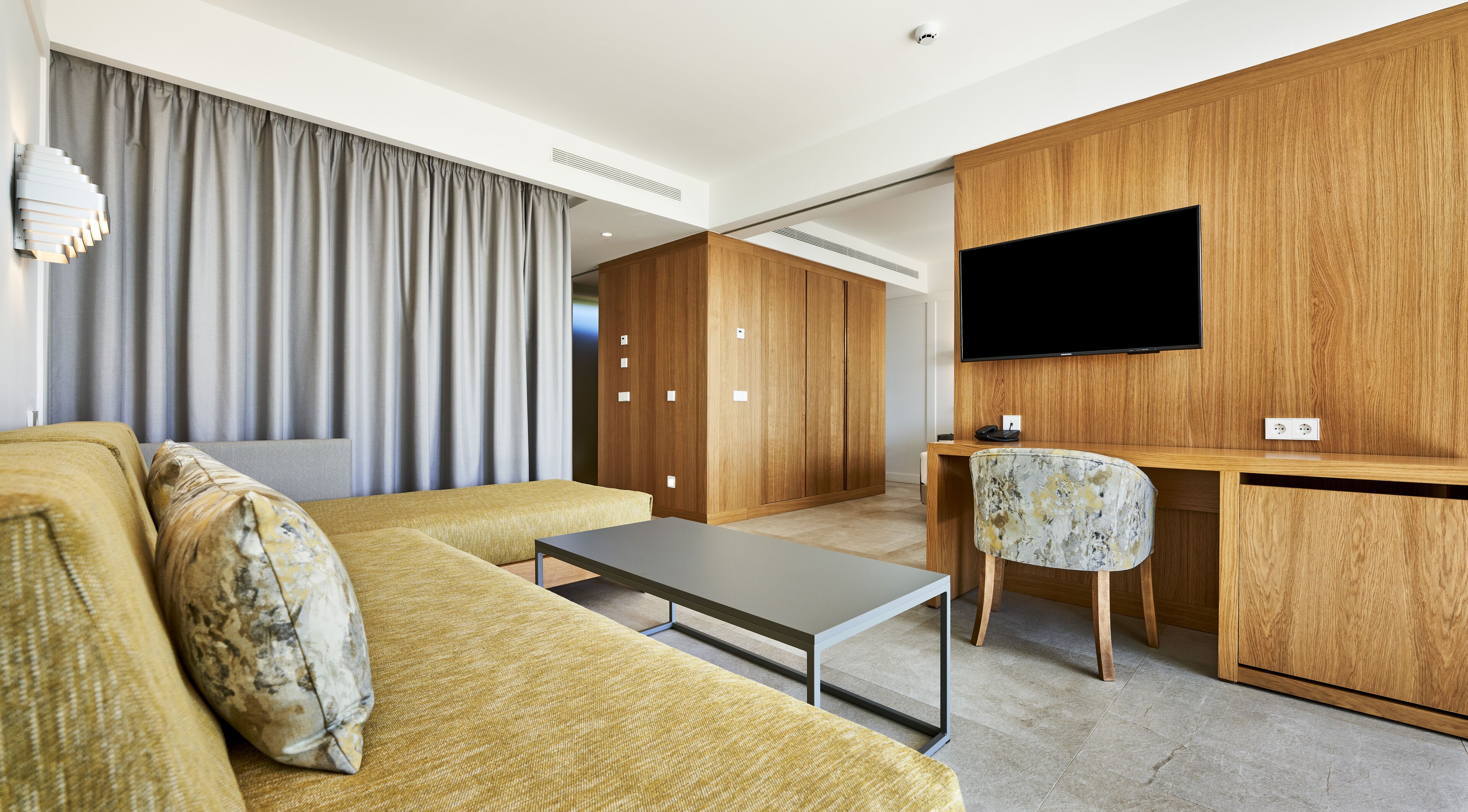 A modern hotel room with a sofa, a small table, a desk with a chair, a television, and a wardrobe.