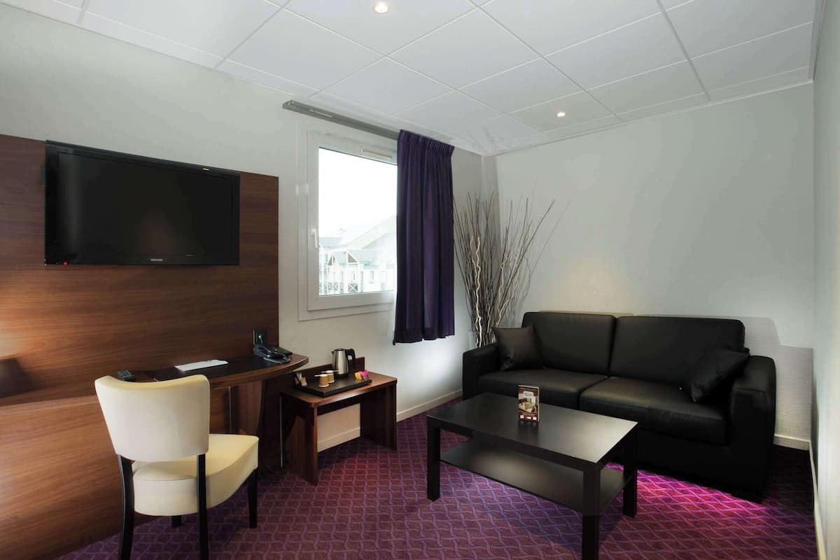 A modern hotel room with a flat-screen TV mounted on the wall, a black leather sofa, a white armchair, a small wooden coffee table, and a window with a view of buildings.