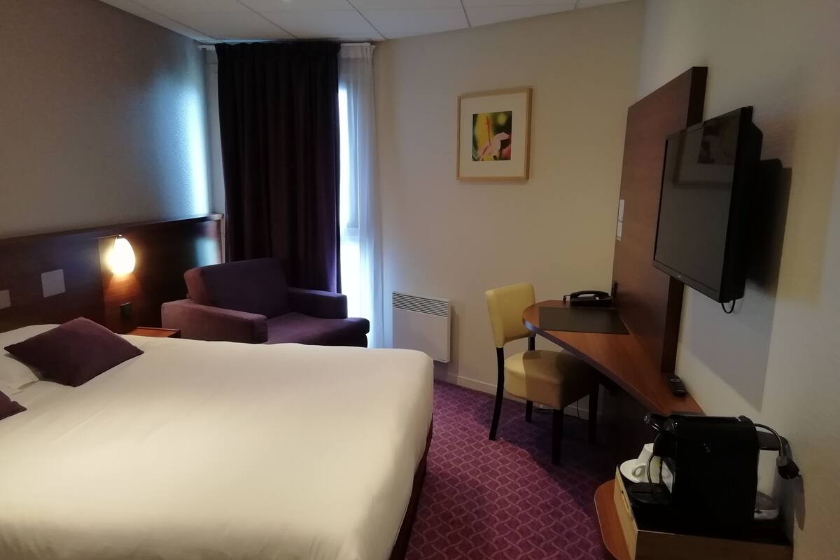 A hotel room with a large bed, a desk, a chair, a TV, and a coffee machine.