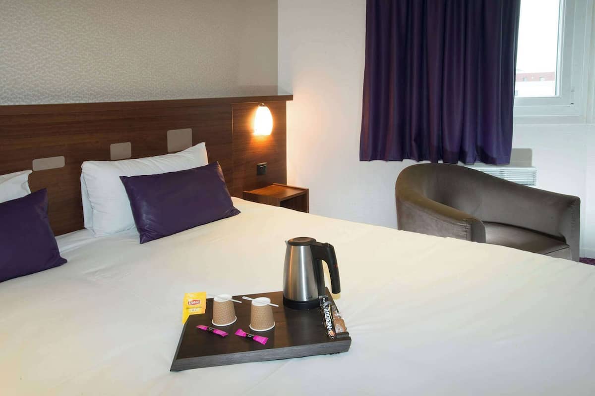 A hotel room with a neatly made bed, a bedside table with a lamp, a chair, and a tray with a kettle, cups, and a snack package.