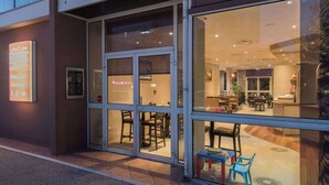 A modern hotel entrance with glass doors, a reception desk, and a seating area visible through the windows.