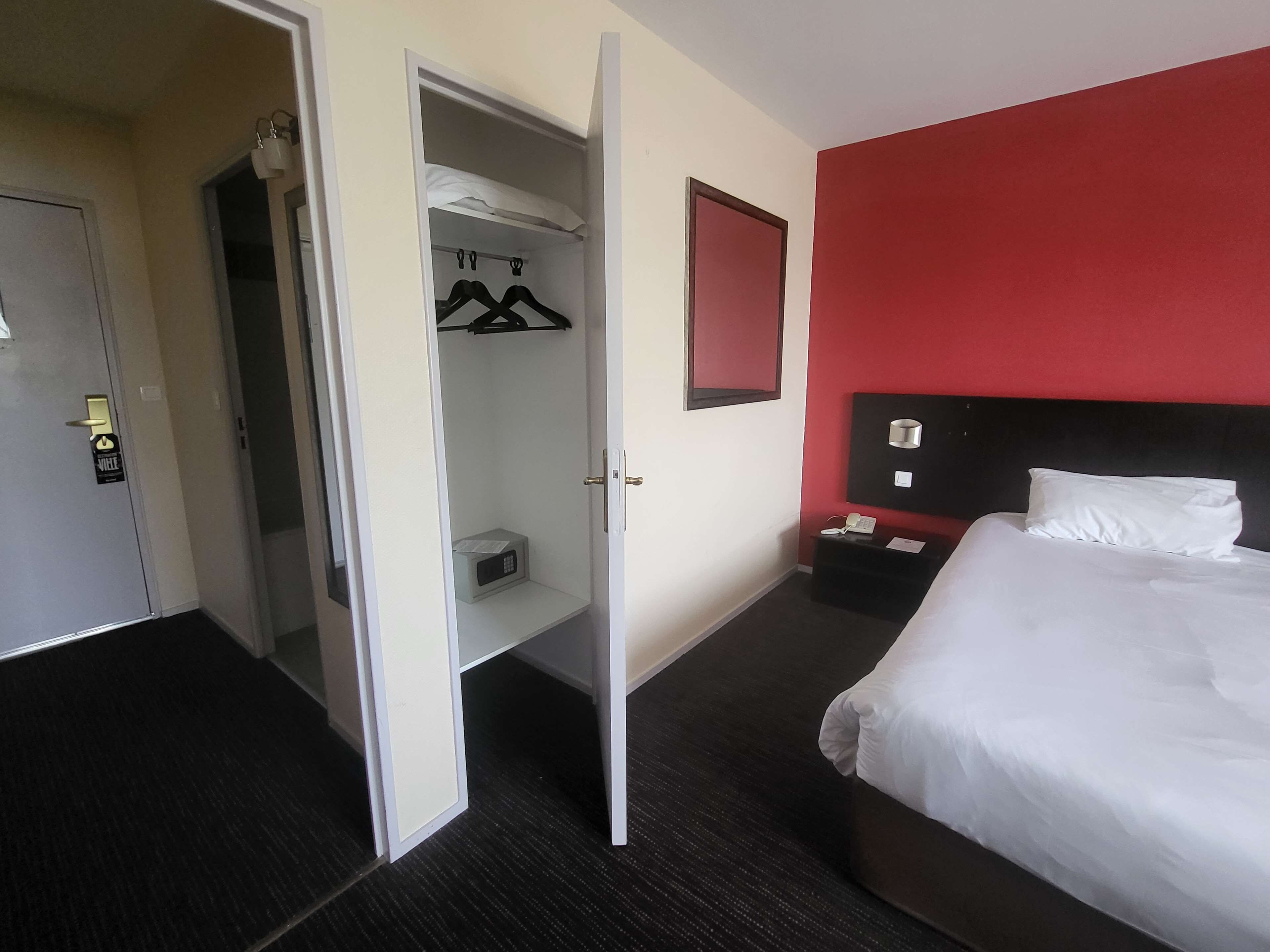 Habitación de hotel con cama, mesita de noche con teléfono, pared de acento roja, armario con perchas y microondas.