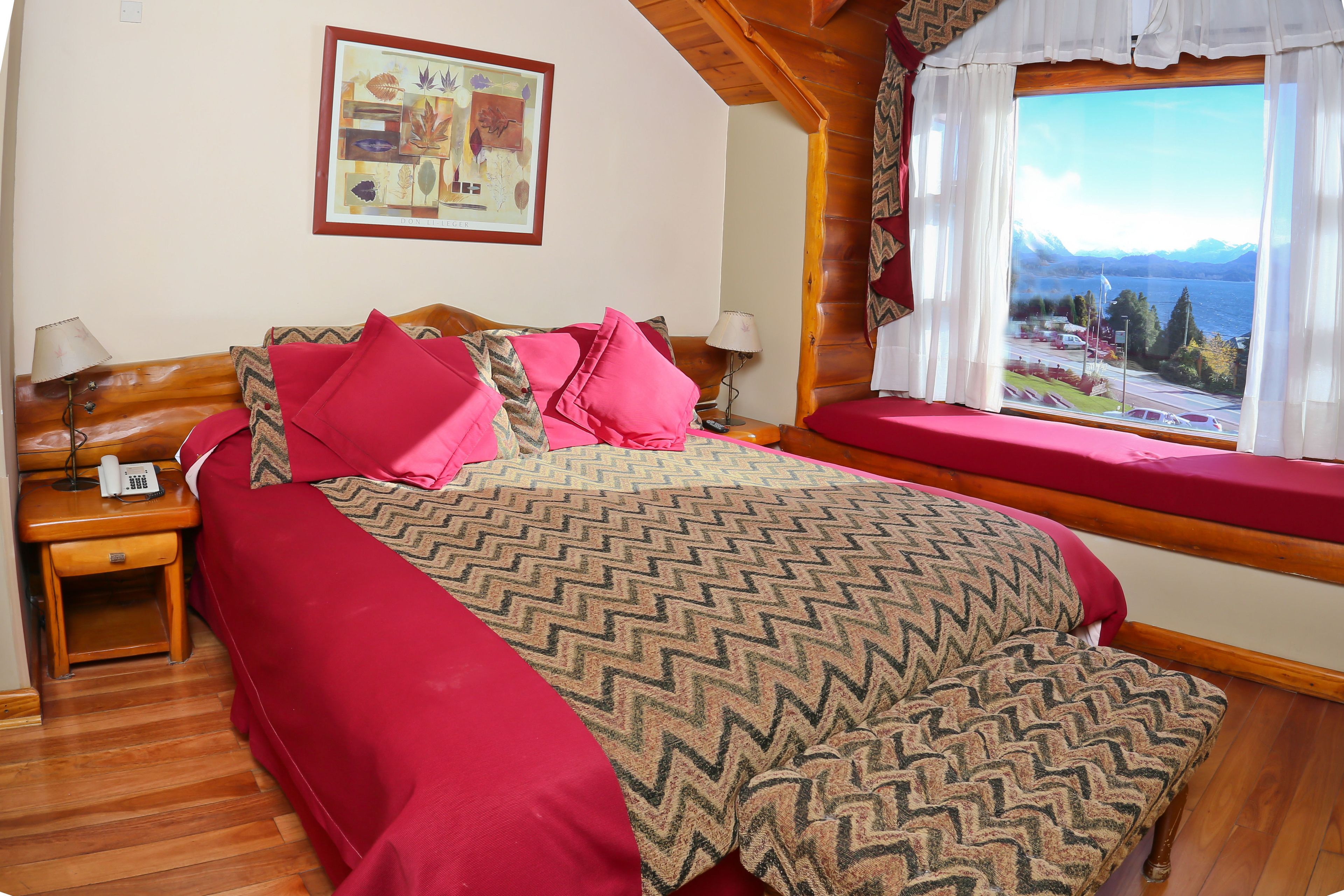 A bedroom with a large bed, two bedside tables, a window with a view of mountains and a lake, and a painting on the wall.