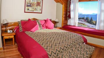 A bedroom with a large bed, two bedside tables, a window with a view of mountains and a lake, and a painting on the wall.