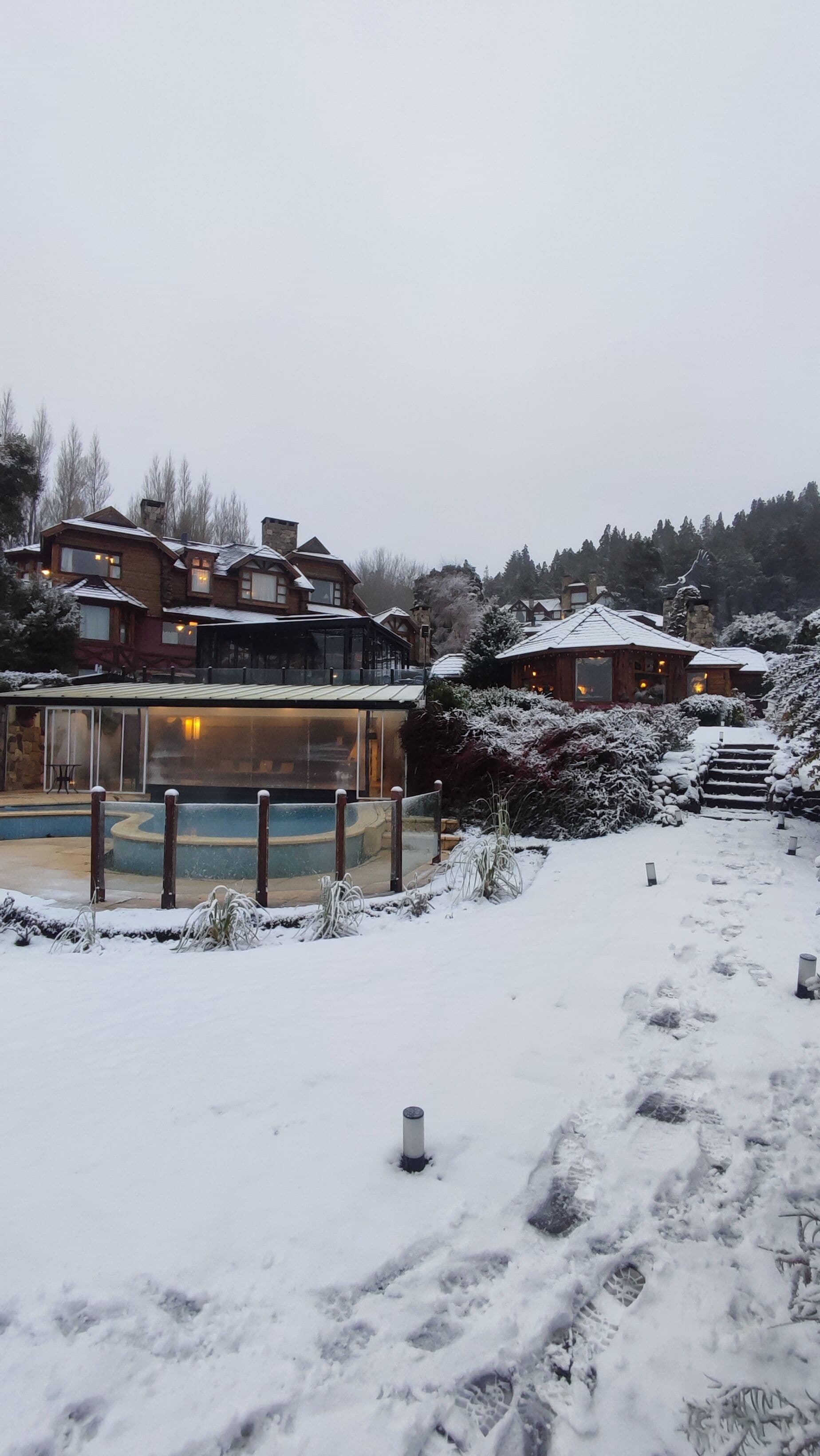 A snow-covered outdoor area with a pool, wooden buildings, and a staircase leading up.