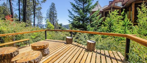 Una terraza de madera con bancos y taburetes hechos con troncos, rodeada de vegetación exuberante y árboles altos.